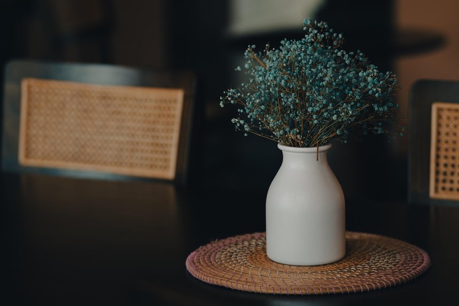 Florero de cerámica con flores secas sobre una mesa de madera, con sillas de ratán al fondo